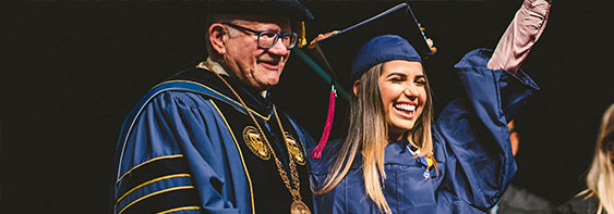 Latin American female student dressed in commencement regalia smiles with her arms raised high during her graduation ceremony