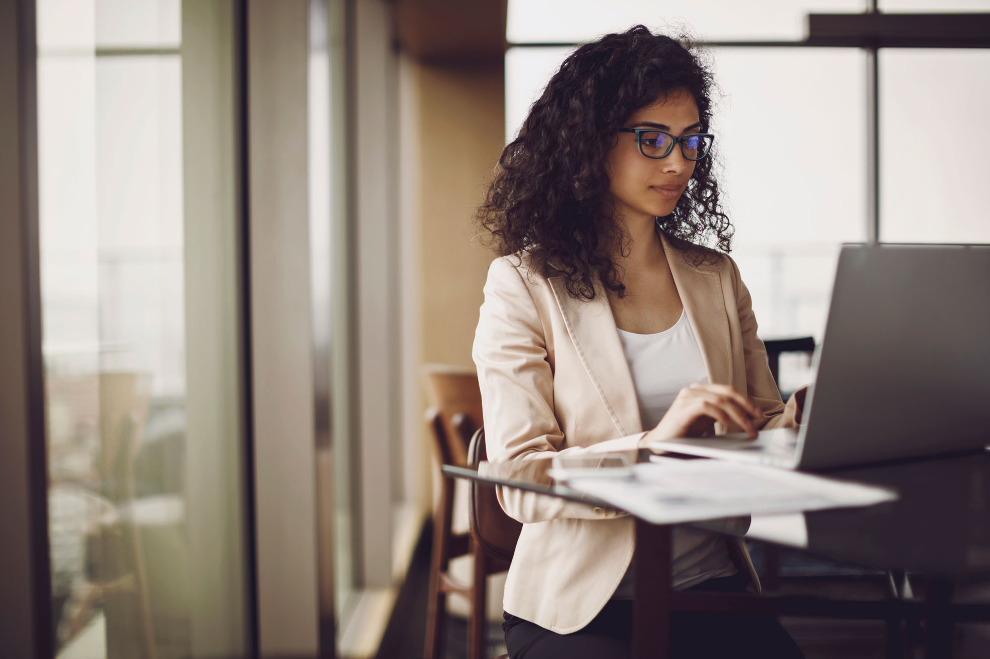 female with dark curly hair and glasses dressed in business attire looking at laptop
