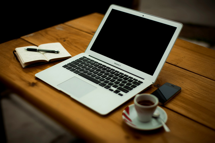 a laptop on a wooden desk beside a small notepad and a cup of coffee